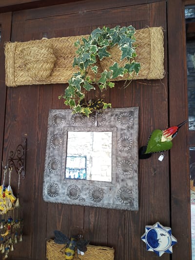 The image shows a rustic wooden door with various decorative items. At the top, a woven straw mat with a raised square section is mounted horizontally. A sprig of ivy with variegated green and white leaves is draped over the mat. Below the mat, a square mirror with a distressed, embossed metal frame is centered on the door. The frame features a repeating pattern of circular motifs resembling sunflowers. To the right of the mirror, a decorative wooden bird with a green leaf-shaped body and red accents is attached to the door. Below the mirror, a metal bee with black and yellow markings rests on a woven basket. To the lower right of the mirror, a blue and white ceramic tile depicting a crescent moon and stars is mounted on the door. A string of bells and colorful beads hangs from a metal hook on the left side of the door, to the left of the mirror. The overall impression is of a quaint shop or entranceway, possibly in Besalú, Spain, given the style of the decorations which evoke a Mediterranean or artisanal aesthetic. The door is made of dark stained wood planks.