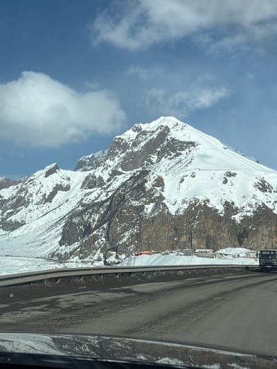 The scene depicts a snow-covered mountain landscape in Almasiani, Georgia. A towering, snow-capped mountain, featuring prominent dark rock faces interspersed with snow, dominates the background under a blue sky with scattered white cumulus clouds. In the foreground, a multi-lane asphalt road, appearing damp and soiled, occupies the lower portion of the frame. A concrete road barrier separates the immediate foreground from the area to its right. A dark-colored commercial truck is visible on the far right of the road, oriented away from the camera. At the base of the mountain, beyond the road, several small, snow-covered structures are present, one of which has a distinct orange-colored roof. The image perspective suggests it was taken from inside a vehicle, evidenced by a slight reflection at the bottom edge. No individuals are depicted.