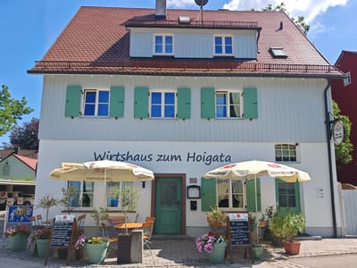 The image shows the exterior of a two-story building, likely a restaurant or beer garden, with the sign "Wirtshaus zum Hoigata" prominently displayed on the white facade. The building features a red tiled roof, white siding, and light blue trim around the windows. Green shutters are closed on some windows and open on others. Two large white umbrellas, branded with "STORCHENBRAU," provide shade for outdoor seating areas. Several potted plants and flower arrangements are placed in front of the building. A green door is centrally located on the ground floor. A blackboard menu is propped up near the entrance. In the background, there are glimpses of other buildings and trees. The scene appears to be set in a town or village under a clear blue sky. The image was taken in Memmingen, Germany. There are no people visible in the image.