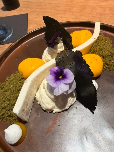 A close-up image displays a meticulously plated dessert presented on a dark, reddish-brown, circular plate. The dessert features an arrangement of white, creamy mounds, each topped with a small, vibrant purple edible flower. Scattered among these are multiple smooth, yellow, spherical components. Dark, irregularly shaped, perforated crisps are vertically integrated into the composition, and a long, curved, off-white strip of an unknown solid substance diagonally traverses the plate. Two distinct piles of green, granular crumble are visible on the left and right sides of the plate, bordering the central arrangement. A smaller yellow sphere, topped with a white meringue kiss, rests in the bottom-left corner. The dessert is situated on a light brown wooden table, with a dark napkin and the partial rim of a clear glass visible in the upper-left background. The scene is located in Kiryat Ono, Israel.