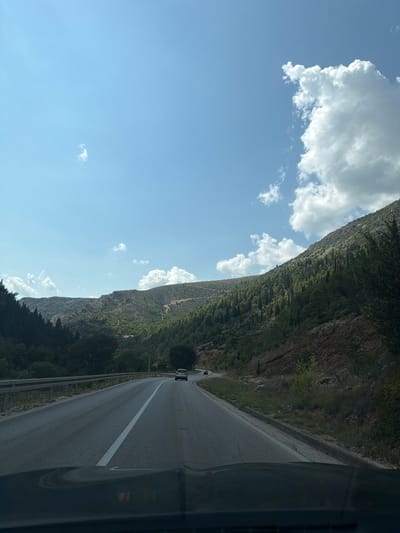 The image depicts a scenic view of a winding asphalt road in a mountainous region, likely near Stolac, Bosnia and Herzegovina. The road, with a white dashed line dividing the lanes, curves through a landscape dominated by lush green hillsides covered in trees and sparse vegetation. On the right side of the road, a steep, rocky incline rises, sparsely vegetated with trees. On the left side, the terrain is also hilly and forested. The sky is a vibrant blue, dotted with fluffy white cumulus clouds. In the distance, a few vehicles are visible on the road, including a white car. The foreground shows the hood of a dark-colored vehicle, indicating the perspective is from a driver or passenger. The overall impression is a serene drive through a natural, rugged environment.
