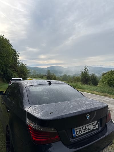 The image shows the rear of a dark-colored BMW sedan parked on the side of a rural road, likely in or near Boltata, Bulgaria, given the license plate format. The car's rear windshield and trunk are wet, with visible raindrops and streaks, indicating recent precipitation. The car is oriented facing away from the viewer and slightly towards the right. A license plate with the text "EB 5627 BH" and the Bulgarian country code "BG" is clearly visible on the trunk.

In the background, a lush green landscape unfolds, characterized by rolling hills covered in trees and vegetation. A light mist or fog hangs in the valleys between the hills, creating a serene and somewhat dramatic atmosphere. The sky is overcast with a mix of grey and white clouds, with hints of sunlight breaking through on the left side of the frame.

To the left of the BMW, another vehicle, possibly a white SUV, is parked on the grass shoulder. The road itself appears to be asphalt and curves gently into the distance. The overall environment suggests a natural, undeveloped area, typical of a scenic route in a mountainous or hilly region. There are no people visible in the image. The scene is static, with no discernible action taking place.