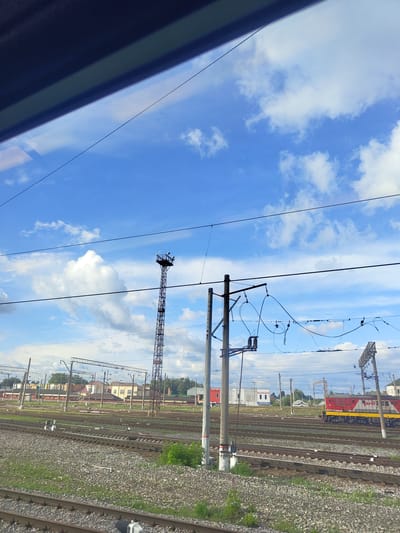 The image shows a view from a train window, looking out at a railway yard under a blue sky with scattered clouds. In the foreground, there are multiple sets of train tracks covered with gravel and some sparse vegetation. Several utility poles with overhead wires are visible throughout the yard, some with complex wiring systems and insulators. A tall, lattice-like tower with lights at the top stands prominently in the middle ground. In the background, there are various buildings, including what appear to be small industrial or service buildings with signs, and some residential-looking structures. A red and yellow train engine is parked on one of the tracks in the distance, to the right of the frame. The overall scene depicts an active railway operational area within a city, likely Фруктовая, Russia, given the context. There are no people clearly visible in the image.
