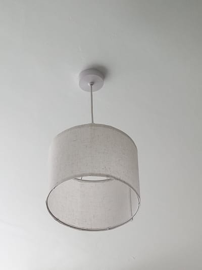 A cylindrical lampshade with a textured, light-colored fabric hangs from a white ceiling. The lampshade is suspended by a thin, white cable attached to a white circular ceiling rose. The background is a plain white ceiling, slightly textured. The lighting suggests it is daytime, and no specific weather or location cues are visible other than the overall indoor setting. No people are present, and no text is visible in the image.