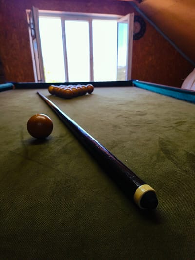 This indoor photograph, captured in Терновский округ, Ukraine, depicts a billiard table situated in a room with a sloped, wood-paneled ceiling and walls. In the foreground, a dark-colored billiard cue stick lies on the worn, olive-green felt surface of the table, positioned beside a single brown billiard ball. Further down the table, a triangular rack contains approximately fifteen brown billiard balls. A large, multi-pane window dominates the background, allowing bright daylight to illuminate the scene, with the leftmost pane appearing slightly ajar. To the right of the window, a dark, circular object is mounted on the wooden wall. No individuals are present in the image.