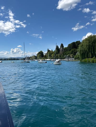 The image depicts a wide shot of a body of water with numerous sailboats and a few motorboats docked or anchored. The water is a vibrant blue-green, with visible ripples and reflections of the sky. In the background, a lush green shoreline is lined with trees, some of which are weeping willows. A large, traditional-looking building with a brown facade is visible amongst the trees on a slight elevation. A diving platform with a wooden structure is also present near the shore. The sky is bright blue with scattered white, fluffy clouds. The foreground shows the dark blue edge of what appears to be a boat or pier. The scene is set in Hünibach, Switzerland, near a waterfront area, likely a marina or harbor on a lake. No people are clearly visible in the image.