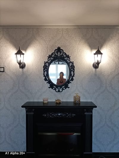 The image depicts an interior scene featuring a black fireplace with decorative molding. Above the fireplace hangs an ornate, oval-shaped black mirror with intricate detailing. On the mantelpiece, there are a few decorative items, including a small gold-colored turtle figurine and a glass perfume bottle. Two wall-mounted lantern-style lights flank the mirror, casting a warm glow on the textured, damask-patterned wallpaper. In the mirror's reflection, a person is visible taking a selfie, holding a phone. The overall ambiance suggests a well-decorated living space or entryway. The location is identified as Улукаўскі сельскі Савет, Belarus.