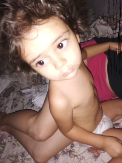 The media file is a close-up, portrait-style photograph of a young child. The child has dark, curly hair and is looking towards the camera with wide, dark eyes. The child appears to be wearing a diaper. The background is blurred, but suggests a domestic setting with patterned bedding. The lighting is dim, hinting at a nighttime or indoor scene. The child's pose is relaxed, with their legs crossed. There are some vertical black lines on the right side of the image, possibly an artifact from the capture or editing process. No text is visible in the image.