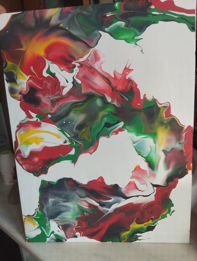 This is a photograph of an abstract acrylic pour painting. The painting features a dynamic flow of colors, primarily red, green, yellow, black, and white, creating swirling patterns and organic shapes across a white canvas. The colors blend and separate in a fluid, marbled effect, with some areas appearing translucent and others opaque. Small white specks are scattered throughout the painted sections, adding a textured detail. The composition is abstract, with no discernible figures, objects, or specific scene. The activity taking place is the visual representation of the paint-pouring process, showcasing the way the colors have interacted and dried. The lighting suggests an indoor setting, possibly a studio or a room where the painting is displayed. There are no visible text elements in the image. The overall impression is one of energetic movement and vibrant color.