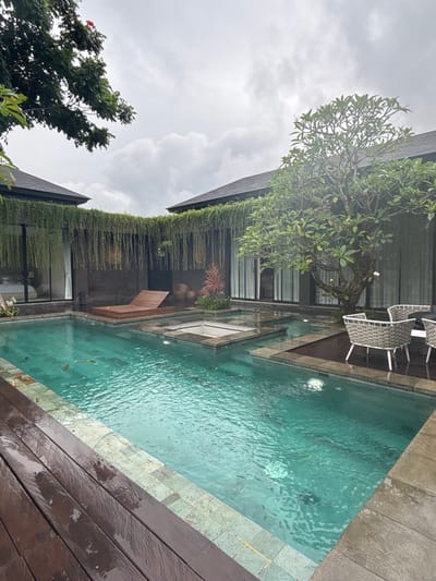 A serene view of a modern house with a long, narrow pool. Lush green plants cascade down the building's exterior. Next to the pool, wooden planks create a deck-like area. White wicker chairs sit near the water, shaded by a tree. The sky is overcast, suggesting a cloudy day.