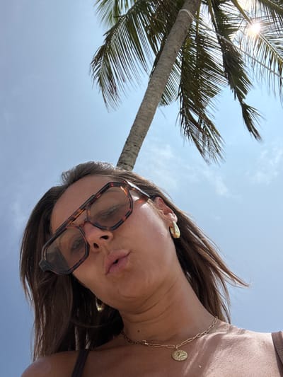 A low-angle shot captures a woman looking down towards the camera, set against a bright, sunny sky and a prominent palm tree. The woman's face is central, adorned with large, tortoiseshell-patterned sunglasses that cover her eyes. Her lips are slightly parted, and her dark brown hair frames her face, appearing wind-swept or styled. She wears a golden-colored, sculptural earring on her visible ear and a delicate gold chain necklace with a circular, coin-like pendant around her neck. A portion of a light brown or beige top is visible on her right shoulder.

In the background, a large palm tree trunk stretches diagonally across the left side of the frame, with its green fronds fanning out against a clear, light blue sky. A sun flare is visible through the palm fronds in the upper left corner, indicating bright daylight conditions. The scene evokes a tropical setting, consistent with the location of Thoddoo, Maldives. The overall activity appears to be a relaxed pose for a photograph, likely a selfie, under pleasant weather conditions. There is no visible text in the image.
