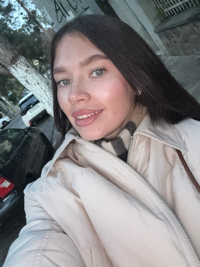 A female individual is captured in a close-up, eye-level selfie-style photograph taken on a street in the Municipal Unit of Elliniko, Greece. The subject, positioned centrally, has long, dark brown hair, light blue eyes, and fair skin, with a faint smile directed at the camera. She wears a light beige collared jacket with a prominent silver zipper, revealing a striped scarf (black and beige) underneath. A small, reflective stud earring is visible in her right ear, and a brown strap, likely from a bag, is visible over her right shoulder.
The background, slightly out of focus, reveals an urban street environment. To the subject's left, a large tree with light-colored, mottled bark stands next to a sidewalk. Parked vehicles are visible on the street behind the tree, including a white SUV/hatchback with a Greek license plate ("YN...") and a dark-colored sedan. To the subject's right and upper right, a stone wall and a white metal fence or railing of a building are partially visible. Overhead, a utility pole or similar structure displays weathered posters and graffiti, including visible Greek characters ("ΑΛ"). The ambient lighting suggests late daylight or early evening conditions.