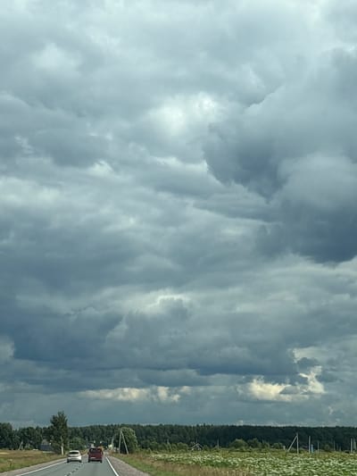 The image depicts a cloudy sky over a rural road in Novaya Pustosh, Russia. Two vehicles are visible on the asphalt road: a white car followed by a red minivan. The road stretches into the distance, bordered by a field of white wildflowers and a dense line of green trees. The sky is filled with dark, heavy clouds, suggesting an overcast or stormy day. The scene is captured from a moving vehicle, as indicated by the perspective.