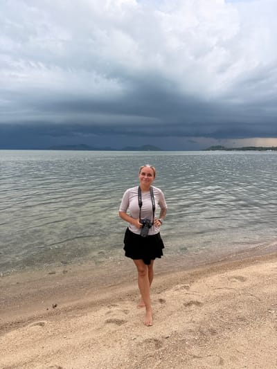 A medium shot captures a woman standing on a sandy beach, facing forward with a camera around her neck. She has light skin, blond hair pulled back, and is wearing a light-colored t-shirt and a black skirt. The camera strap is visible, with the camera body resting against her stomach. Her left hand is on her hip, and her right hand is near the camera. The background consists of the sea with gentle ripples and a cloudy, dark sky, suggesting an impending storm. In the distance, a hazy shoreline with hills is visible. The sand in the foreground shows footprints and a small crab. The location is identified as Ban Bang Khao, Thailand.