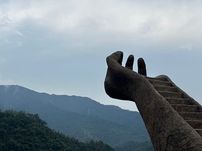 The image shows a large stone sculpture of a hand in the foreground, with stairs leading up the palm and fingers. The sculpture is positioned on a hill overlooking a valley filled with lush green mountains. The sky is overcast with a few wispy clouds. The sculpture is part of the Tianmen Mountain Scenic Area in Qingyuan City, China.