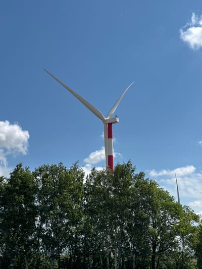 The image shows a wind turbine against a bright blue sky with scattered white clouds. The wind turbine has white blades and a red and white striped tower. In the foreground, there is a dense line of green trees. In the background, another wind turbine can be seen, partially obscured by trees and clouds. The scene appears to be an outdoor landscape in Krivoluch'e-Ivanovka, Russia.