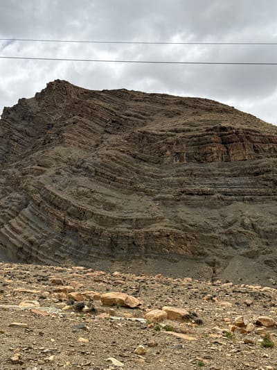 The image shows a close-up of a layered rock formation, possibly a cliff or mountainside, in Zagor town, China. The rock is composed of sediment laid down over time, with distinct strata that show folding and distortion. The ground adjacent to the rock face is covered in loose gravel, stones, and sparse vegetation. In the background, there are power lines against an overcast sky with gray clouds.
