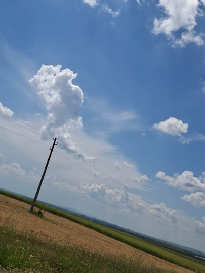 This image depicts an agricultural landscape under a bright, partly cloudy blue sky, captured in the vicinity of Kukorevo, Bulgaria. The scene is presented with an oblique horizon line, sloping upwards from the lower left to the upper right.

The sky, occupying approximately the upper two-thirds of the frame, is clear blue with scattered white cumulus clouds of varying sizes. A large, distinct, irregularly shaped cumulus cloud formation is prominent in the upper left quadrant.

In the mid-left, a dark brown utility pole stands, featuring insulators and at least four parallel power lines extending horizontally across the mid-ground. The terrain immediately below the power lines and pole consists of cultivated land. The foreground and mid-ground on the left and center are dominated by a field of mature, golden-brown grain, indicative of wheat or a similar cereal crop nearing harvest. A small section of green grassy vegetation with a few red poppy flowers is visible at the very bottom left, adjacent to the golden field.

Further in the background, a distinct band of vibrant green agricultural field stretches horizontally across the mid-right. The distant horizon is hazy, revealing faint topographical features consistent with hills or mountains, and several indistinct industrial structures, possibly smokestacks or towers, indicating a distant settlement or industrial area. No individuals, vehicles, or animals are present in the visible area.
