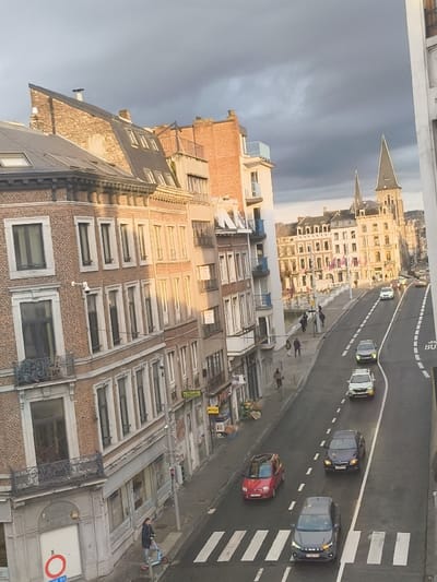 This image captures an elevated view of a city street in Liège, Belgium, specifically along the **Quai sur Meuse** or an adjacent street such as **Rue Léopold**, looking west-southwest towards the **Collégiale Saint-Denis** and the city center.
The scene depicts an urban environment under a partly cloudy sky, with dark, heavy clouds dominating the upper portion and patches of sunlight illuminating specific areas. A multi-lane asphalt road runs diagonally through the center, displaying white painted lane dividers and pedestrian crosswalks. The road surface exhibits wet patches, reflecting light. Vehicle traffic flows in both directions; a dark gray hatchback, a dark blue/purple hatchback, a white SUV, and a dark sedan are visible in the lane approaching the viewer. A red compact car is in the opposing lane. Several additional vehicles are present further down the street. A partially visible "BUS" lane marking is on the far right.
On the left side of the street, a row of multi-story buildings with brick and stucco facades features numerous windows, some with dark frames, and wrought-iron balconies. Ground-floor establishments have awnings and merchandise displays, with a visible sign reading "L'Escale." A white surveillance camera is mounted on one building. At the bottom left, a pole displays a red circular traffic sign with a white bar. A lone individual rides a light blue electric scooter on the sidewalk adjacent to a pedestrian crossing. Further along this sidewalk and across the street along the riverfront, several other pedestrians are walking.
To the right of the street, a body of water, likely the Meuse River, is partially visible, bordered by a low wall. Beyond the river, another line of multi-story buildings, predominantly light-colored, features flags or banners. In the background, the distinctive spires of the Collégiale Saint-Denis rise above the city's skyline, characterized by its tall, octagonal main spire. Distant hills covered with trees are visible on the horizon. The sunlight casts long shadows, suggesting late afternoon or early morning.