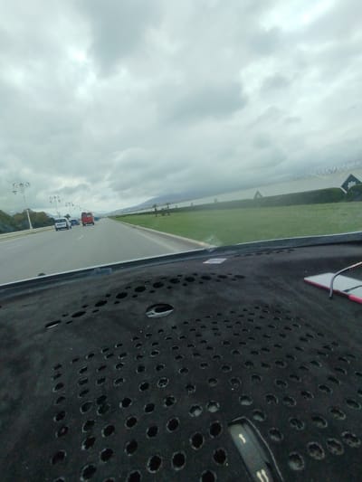 The image shows a view from inside a car, looking out onto a multi-lane road. Several vehicles, including a red van and other cars, are visible ahead on the road. A grassy area borders the road on the right side, with a line of greenery and a distant body of water or coastline beyond. The sky is overcast with gray clouds. The dashboard of the car, which has a perforated design, is prominent in the foreground, with a smartphone resting on it. This scene appears to be located in the city of Tétouan تطوان, Morocco, based on the architectural style of the street lamps and the general landscape.