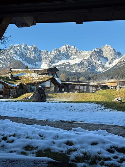 The image depicts a panoramic view of a distinctive architectural complex set against a backdrop of snow-capped mountains in Going am Wilden Kaiser, Austria. The scene is observed from under a dark, wooden overhang.
In the foreground, patches of melting snow are scattered across green grass, revealing underlying vegetation. A partially snow-covered path or road traverses the lower mid-ground.
The mid-ground features a series of low-profile buildings characterized by extensive green, grassy roofs, interspersed with sections of wooden shingles and multiple skylights. The structures incorporate natural stone cladding and large, angular windows that reflect the bright blue sky. A prominent, dark natural boulder is positioned in front of one of the buildings. To its right, a plume of steam or mist rises from the ground, possibly indicating a geothermal or heated water feature. Small, dark, indistinct figures, potentially waterfowl, are present near the steam.
The background is dominated by a clear, deep blue sky above a majestic range of snow-covered mountains with rugged, exposed rock peaks. The mountains appear sharply defined under bright sunlight. The overall environment suggests a crisp, sunny day with remnants of winter snow. No explicit human figures or interactions are discernibly present.