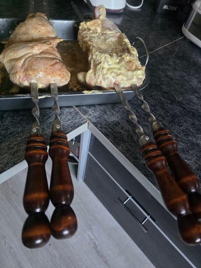 This media file shows two whole chickens prepared for roasting, each impaled on a metal skewer with wooden handles. The chickens are coated in a yellow marinade, with visible flecks of herbs and spices. One chicken is positioned above the other, both resting in a metal tray. The scene appears to be indoors, likely a kitchen, with a dark, textured countertop in the background and a light gray wooden floor. To the right, a portion of a dark gray cabinet with a silver handle is visible. The lighting suggests an indoor setting, possibly during the day, but no specific time or weather cues are discernible. No people are visible. The image is shot from a top-down perspective, with the skewers angled upwards. The overall impression is one of food preparation.