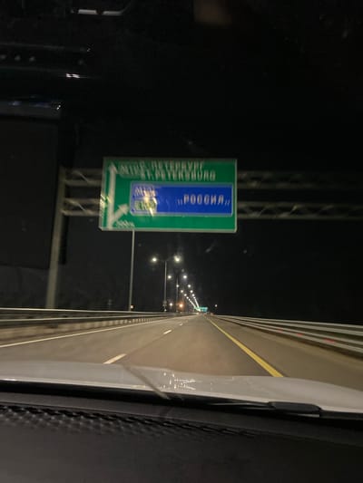 The image shows a nighttime view from inside a vehicle driving on a highway. A green road sign with white lettering is visible, indicating directions to Saint Petersburg and Russia. The sign has a blue rectangle with text in Russian. The highway has multiple lanes separated by a yellow line and guardrails on the right. Streetlights illuminate the road ahead. The overall scene suggests travel on a highway at night. The location is identified as Kolomna, Russia.