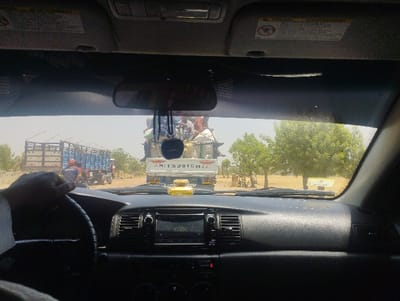 The image shows the view from the driver's seat of a car. The driver is holding the steering wheel with their right hand. The driver is facing forward, looking out the windshield. The car is stopped behind a white Mitsubishi truck, loaded with various goods and materials,  which appears to be stopped on the side of the road. The driver's car is facing a blue, flatbed, enclosed cargo truck, also parked on the side of the road. The cab of the truck is visible, and there is one person inside. There is another person standing near the truck on the driver's side. Both trucks appear to be parked in a narrow, dusty road. The road is lined with trees and foliage. The image appears to be taken in Bajoga, Nigeria (latitude: 10.864, longitude: 11.434). The sky is bright and clear. The driver's dashboard is visible. The driver's side sun visor is down and a warning sign can be seen.  A rearview mirror is visible in the upper left-hand corner of the image. There are several small items visible in the backseat. The image is taken from the perspective of the driver.