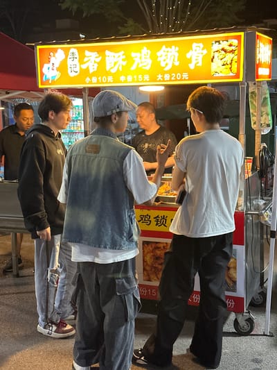 An outdoor street food stall in Eshan Yi Autonomous County, China, is illuminated by a bright yellow sign that reads "李记香酥鸡锁骨" (Li Ji Crispy Chicken Ribs) and displays prices for different portion sizes. Four individuals are present. Two young men, one wearing a black hoodie and grey pants, the other in a denim vest over a white t-shirt and grey cargo pants, are standing in the foreground facing the stall. A vendor, an adult male with short hair, is visible behind the counter of the food stall, appearing to interact with one of the young men. Another adult male in a dark shirt is standing to the left of the stall. The stall is stocked with food items, and bottles of beverages are visible inside. The overall setting suggests a nighttime scene with artificial lighting.