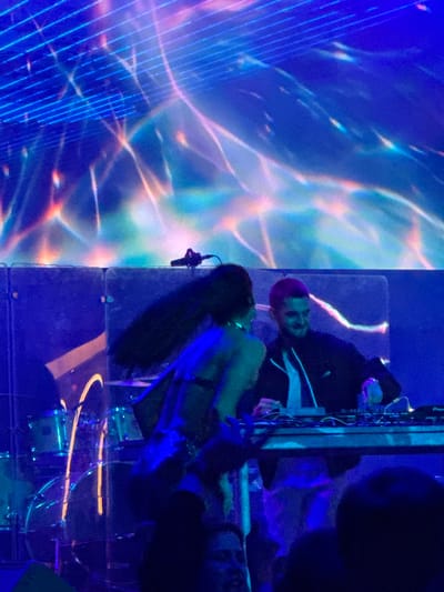 An undated, indoor, medium shot captures a DJ set at a night event, possibly a concert or club, with a dynamic blue and pink light display projected onto a screen in the background. The foreground shows a DJ with short, reddish-pink hair, wearing a black jacket over a white shirt, actively engaged with a DJ console. To his left, a woman with long, dark hair is dancing, wearing a shimmering, dark, outfit. The scene appears to be set in Akhalubani, Georgia, based on the provided context.