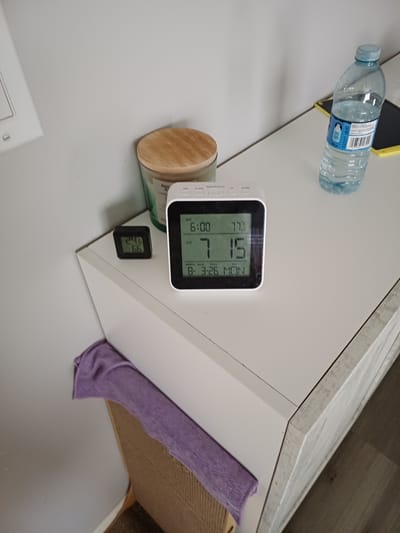 In Morden, Canada, a white nightstand displays various objects. A white digital clock with a black frame shows the time as 6:00 AM, the temperature as 77.3 degrees Fahrenheit, and the date as March 26th, Monday. Next to it, a smaller black digital thermometer reads 24 degrees Celsius and 66% humidity. To the left, a glass jar candle with a wooden lid sits on the nightstand. A clear plastic water bottle with a blue cap and a blue label is positioned on the right side of the nightstand. A folded purple cloth is draped over the edge of the nightstand. The background consists of a plain white wall.