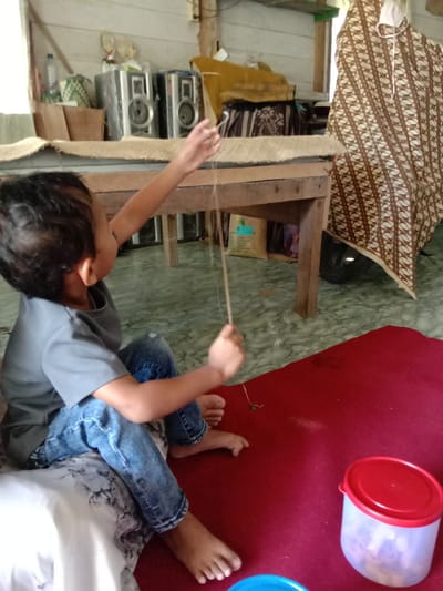 A child sits on a white pillow on a red rug in a room in Blangkarieng, Indonesia. The child is wearing a grey shirt and blue jeans. The child is holding a string. A red and clear plastic container and a blue container are on the rug near the child. Behind the child is a wooden table with two stereo speakers and other items on top. A brown and white patterned fabric covers an object behind the table. The floor is tiled with a marbled pattern.