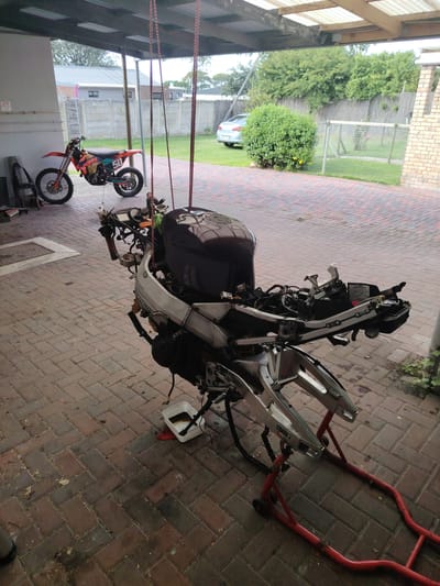 The image depicts an outdoor workshop area in Port Elizabeth, South Africa. The ground is paved with red bricks in a herringbone pattern. A deconstructed motorcycle is suspended from above by red ropes. The motorcycle's fuel tank is present, but much of the rest of the bike is dismantled. A smaller dirt bike with orange and teal accents is parked in the background near a white garage wall, and motorcycle tires are also visible nearby. A car is parked in the background near a green lawn and fencing. A white plastic container sits beneath the suspended motorcycle, likely collecting fluids. The scene suggests ongoing motorcycle repair or maintenance.