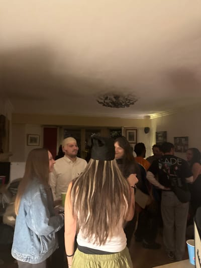 Several people stand in a room. Some face the camera, while others are partially obscured. There's a man in a white shirt and head covering, and a woman with long, highlighted hair. Others wear casual clothing like jeans and t-shirts. A decorative piece is on the wall near the ceiling. The room is lit by warm, indirect light, with multiple framed pictures on the back wall.