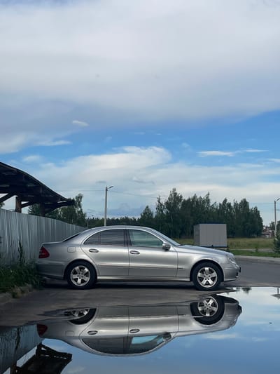 A silver sedan is parked on asphalt next to a corrugated metal fence under a cloudy blue sky. The car's reflection is visible in a large puddle of water on the ground in front of it. In the background, there is a row of trees, a light pole, and a small building. The scene is located in the city of Sosnovka, Russia. There are no people or animals visible in the image.