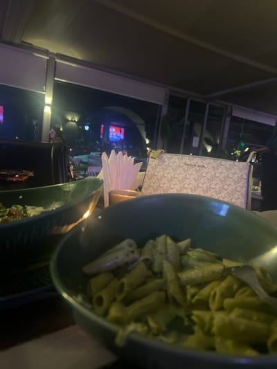 A close-up shot showcases a bowl of penne pasta coated in a creamy pesto sauce. Adjacent to the pasta, a folded stack of white paper napkins stands neatly. The background reveals a softly illuminated restaurant interior with other tables, a purse, and a window, providing a glimpse into the outdoor nightscape. The composition of the photograph is close and intimate, drawing focus to the pasta.