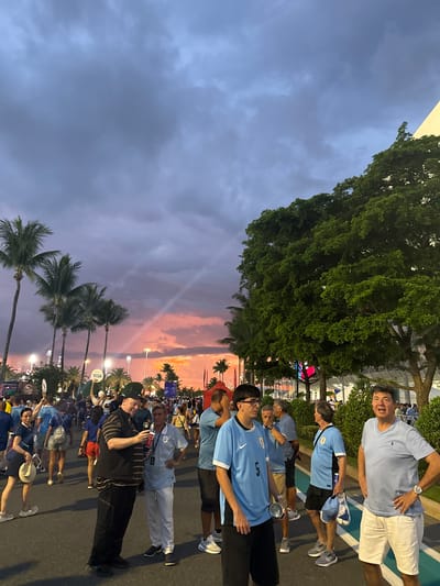 In Miami Gardens, United States of America, on a paved street, a crowd of people are gathered under a dramatic sunset sky. The sky is filled with dark, heavy clouds, interspersed with streaks of pink and orange light, creating a dramatic and colorful atmosphere. Several palm trees are visible in the background, their fronds silhouetted against the sky.
The people in the foreground are mostly men, many wearing light blue t-shirts with national team insignia, suggesting they might be attending a sporting event. Some are engaged in conversation, while others are looking at their phones or holding drinks. One man in the center is wearing a black and brown striped shirt and black pants, holding a phone and a red cup. To his right, another man in a light blue shirt and white pants is standing with his hands on his hips, looking towards the viewer.
Further in the background, more people are visible, along with tents, lights, and fences, indicating a public gathering or event venue. The overall scene captures a moment of anticipation and community as people gather outdoors in the evening.