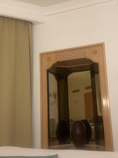An ornate, rectangular, light-brown wooden mirror hangs against a white wall. The mirror reflects a room, revealing a door, a decorative fixture, and two elongated, dark-brown objects resembling pots. Tan-colored curtains drape below the mirror's lower edge. The frame's left side features a lattice pattern with carved rosette details at the corners. A portion of a white molding and light-blue fabric can be seen along the image's left and right edges, respectively.