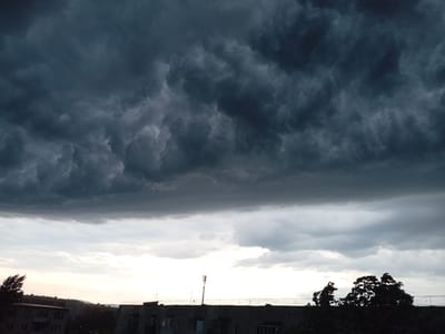 The image depicts a dramatic sky filled with dark, heavy storm clouds looming over the city of Kyshtym, Russia. The clouds occupy the upper two-thirds of the frame, their textured surfaces suggesting an impending downpour. Below the storm clouds, a lighter band of sky breaks through, revealing the softer hues of twilight. In the foreground, the silhouette of buildings and trees is visible against this lighter sky. The architectural style of the buildings suggests Soviet-era construction, a common feature in many Russian cities. A cell tower stands prominently on the rooftop of one building, its antenna reaching towards the turbulent sky. The overall impression is one of atmospheric intensity and the quiet presence of urban life beneath a powerful natural display.