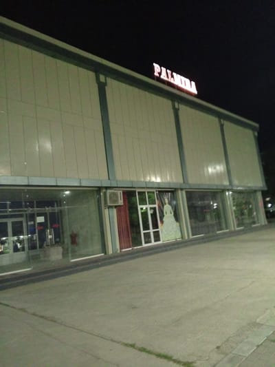 The image shows the exterior of a building at night, identified as the "PALMIRA" venue in Dustlik, Uzbekistan. The building has a modern facade with large glass windows on the lower level, revealing displays inside. A prominent neon sign spelling "PALMIRA" is illuminated on the upper part of the building. Inside the glass-fronted area, a mannequin dressed in a formal gown is visible. The surrounding area appears to be a paved outdoor space, likely a sidewalk or parking lot, with some sparse vegetation. The lighting suggests it is nighttime, with artificial lights illuminating the building and its sign.