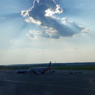 The image shows an airport tarmac under a partly cloudy sky with sunbeams breaking through the clouds. In the foreground, two airplanes are parked. One airplane, with the text "РОССИЯ" visible, is red and white and has a set of stairs attached. Another airplane, lighter in color, is parked further back, also with stairs. Several service vehicles are present on the tarmac, including a fuel truck near the lighter-colored plane. In the distance, a green field and a treeline can be seen, with industrial structures and buildings on the horizon. The scene appears to be taking place at the Naberezhnye Chelny Airport in Naberezhnye Chelny, Russia.