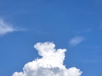 A bright blue sky with scattered clouds. A large, fluffy cumulus cloud dominates the lower center of the frame, with wisps of cirrus clouds stretching across the upper left. A faint contrail from an airplane is visible in the upper right quadrant. The scene appears to be a clear day. No specific location in Rzeszów, Poland can be determined from this image.