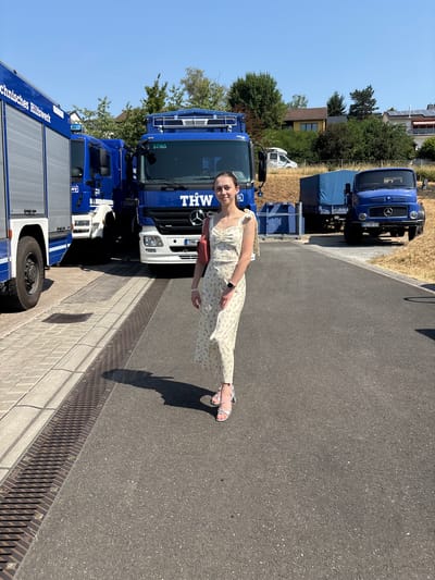 A young woman stands on an asphalt surface in front of several blue trucks. The trucks have "THW" printed on them, indicating they belong to the Technisches Hilfswerk, a German federal agency for technical relief. The woman is wearing a light-colored, floral-patterned dress and silver heeled sandals. She is smiling and looking towards the camera. Behind the trucks, there are trees, houses, and a clear blue sky. The scene appears to be outdoors on a sunny day. The location is Obernburg am Main, Germany, specifically at the THW facility.