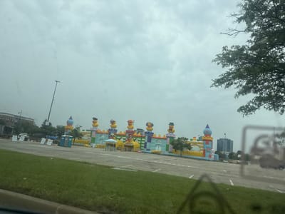 The image depicts an outdoor scene in Schaumburg, Illinois, USA, featuring a large inflatable amusement structure, likely a bounce house or carnival attraction, set up in a paved outdoor area, possibly a parking lot. The inflatable is designed to resemble a castle with multiple turrets and is adorned with several large inflatable duck figures wearing various hats and accessories, such as a police hat, a top hat, and headphones. The inflatable also features large inflatable duck heads with sunglasses as entrances.
In the background, a cityscape is visible with modern buildings, including a tall, dark-colored office building. Trees and utility poles are also present. In the foreground, a grassy area and a paved surface with parking lot markings are visible. Several portable toilets and a blue canopy tent are situated to the left of the inflatable attraction. The sky is overcast, suggesting a cloudy or possibly rainy day. The overall impression is of a temporary event or festival setup.