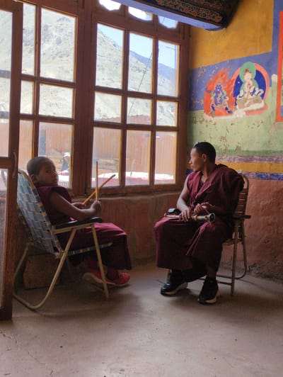 Two individuals, likely monks, are seated in chairs inside a room with a large, multi-paned window. The individual on the left, a younger person, is seated in a blue and white checkered folding chair and is wearing a maroon robe. They are holding two wooden sticks in their hands. The individual on the right, an adult, is seated in a wooden chair and is also wearing a maroon robe. They are holding a decorative object that appears to be a ritualistic implement in their hands. The room has textured, painted walls, with a mural depicting figures on the right wall. The window looks out onto a mountainous landscape. The floor is concrete. The scene appears to be within a monastery or religious building in Pibiting, India.