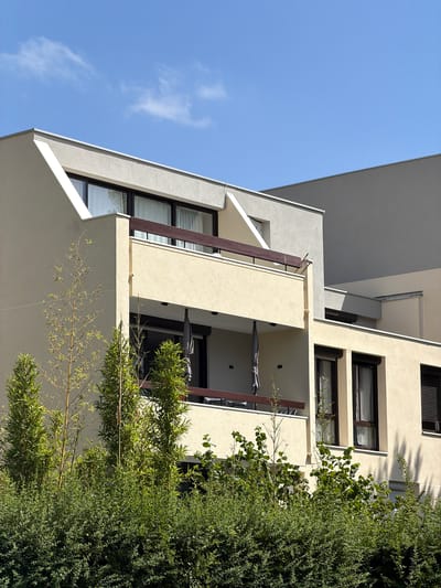 This is a low-angle, vertical shot of a modern building with balconies and large windows, partially obscured by lush green foliage. The building's facade is a mix of light cream and grey concrete. Several balconies are visible, some with folded grey umbrellas. The windows are dark-framed and reflect the bright blue sky and some white clouds. Thin, wooden beams run vertically along some of the balcony edges. Thin branches with small green leaves, possibly bamboo, extend from the right side of the frame and trail across the lower part of the building. The overall impression is of a contemporary residential building integrated with nature. The scene is illuminated by bright sunlight, suggesting it is daytime with clear weather. No people or text are visible. The context provided places the location in Évry-Courcouronnes, France.