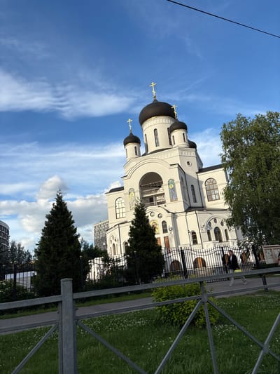 The image shows a white church with black domes topped with golden crosses. The church features arched windows, decorative mosaic panels, and a large arched entryway. In front of the church, there is a black metal fence. Beyond the fence, a paved walkway runs alongside a grassy area dotted with small white flowers. Two dark green evergreen trees are visible to the left of the church, and a tall leafy tree stands to its right. In the background, modern buildings rise above the trees. A person is walking along the paved walkway in front of the fence. The sky is blue with scattered white clouds.