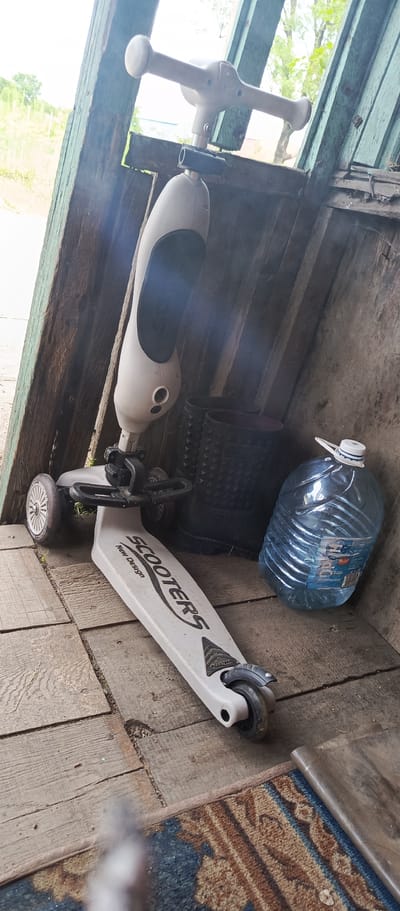 The image depicts a white scooter leaning against a weathered wooden structure. The scooter has a light-colored body with black accents and the word "SCOOTERS" in black lettering on its deck, along with "New Design". It has three wheels, with the front wheel section showing a handlebar attachment. To the right of the scooter, there are two black, spiky boots and a large, clear plastic bottle filled with blue liquid, likely water, with a white and blue cap. The objects are situated on a wooden floor, with a portion of a patterned rug visible in the foreground. The background shows wooden planks and an overcast sky with hints of greenery. The scene appears to be an outdoor storage area, possibly a shed or lean-to, given the rustic construction and the presence of various items. The location is identified as Pogranichny, Russia.