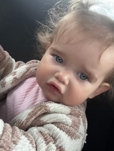 A close-up, head-and-shoulders shot of a baby girl with blonde hair and bright blue eyes is featured. She is wearing a pink ribbed shirt and is partially wrapped in a fuzzy blanket with a pink and white striped pattern. The baby's head is tilted, and her gaze is directed slightly off-camera. The background is dark and indistinct, suggesting an indoor setting. There is no discernible information indicating a location in Braunfels, Germany.