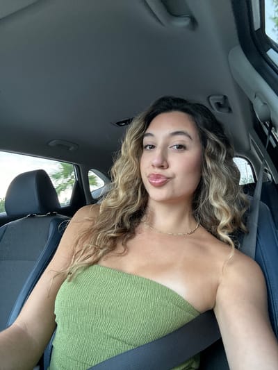 A young woman with long, wavy blonde hair is sitting in the driver's seat of a car, looking at the camera. She is wearing a green, strapless top and a gold necklace. Her lips are pursed and she is wearing lip gloss. The interior of the car is visible, including the headrest of the passenger seat, the ceiling of the car, and a seatbelt. Outside the car window, palm trees and a cloudy sky are visible, suggesting the location is in a tropical or subtropical region, consistent with Hillsboro Beach, Florida, United States. The lighting suggests it is daytime.